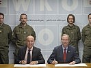 Klug und Leitl beim Unterzeichnen der Kooperations-Vereinbarung, dahinter Milizsoldaten.